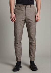 Ljusbruna, texturerade byxor med smal passform, knäppning och sidofickor, i kombination med svarta skor mot en neutral bakgrund.