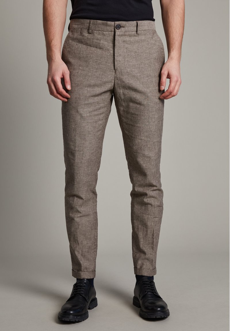 Ljusbruna, texturerade byxor med smal passform, knäppning och sidofickor, i kombination med svarta skor mot en neutral bakgrund.