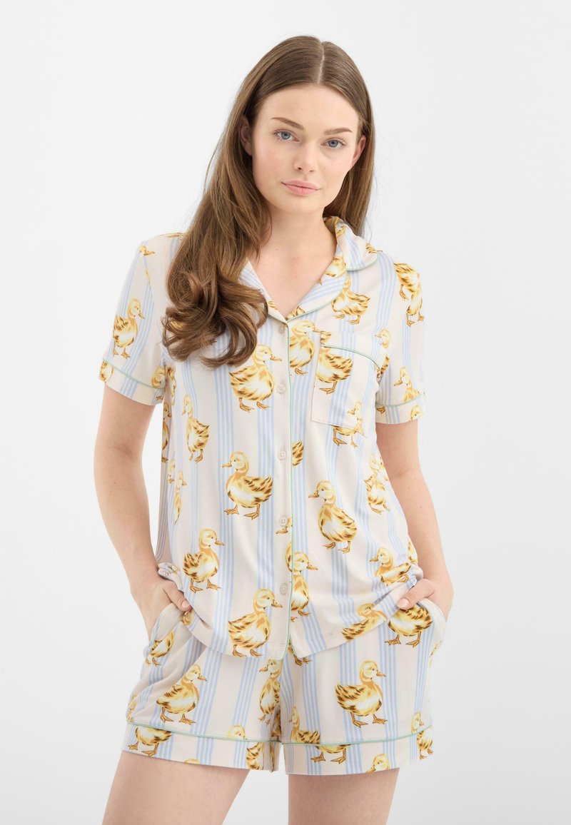 Woman wearing light-colored short-sleeve button-up shirt and shorts with yellow duck print and vertical blue stripes, hands in pockets.