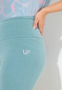 Primo piano di una persona che indossa leggings azzurri chiari UPSports con la mano appoggiata sull'anca, su uno sfondo bianco.