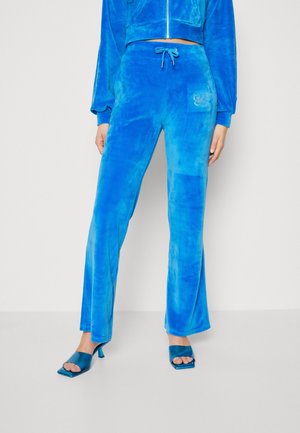 Woman wearing bright blue velour pants with a drawstring waist and crystal butterfly detail on the pocket, paired with matching blue heeled sandals.