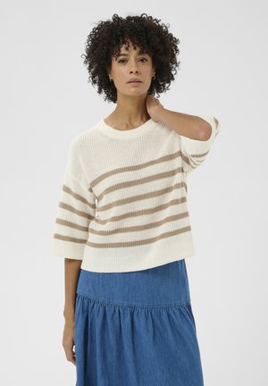 Göndör hajú nő krémszínű, bézs csíkos pulóverben és kék, rakott szoknyában, egy kézzel a nyakát érintve áll.