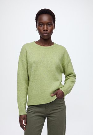 Kvinde med kort hår iført lys grøn sweater og olivengrønne bukser står med den ene hånd i lommen mod en hvid baggrund.