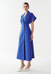 Mėlynas iki kelių ilgio suknelė su trumpomis rankovėmis, mygtukų priekine dalimi ir šoninėmis kišenėmis, pagaminta iš tekstūruoto audinio. Suderinta su baltomis aukštakulnėmis šlepetėmis.