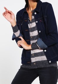 Giacca di denim blu scuro con chiusura a bottoni, tasche frontali e polsini a contrasto blu chiaro, indossata su un maglione grigio a righe.