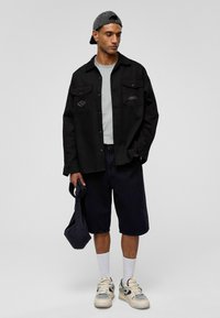 Zwart overhemd met knopen en twee voorzakken, grijs t-shirt, donkere denim shorts, witte sokken en beige sneakers; grijze pet en zwarte tas.