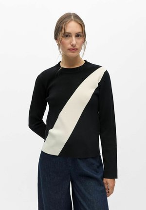 Femme aux cheveux châtain clair portant un pull noir avec une bande diagonale blanche, associée à un pantalon bleu foncé, debout sur un fond blanc.