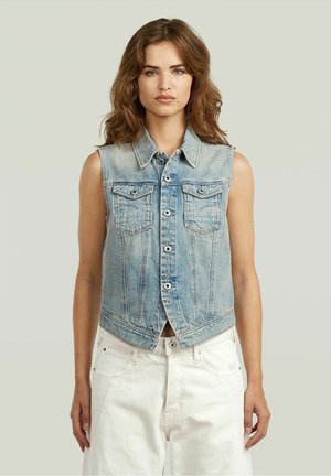 Vrouw met golvend bruin haar, gekleed in een lichtblauw spijkervest zonder mouwen en een witte broek, staand tegen een effen lichte achtergrond.