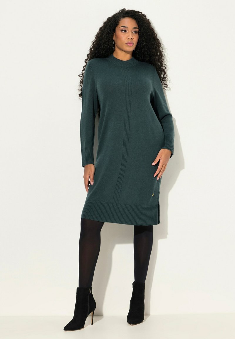 Robe tricotée vert foncé avec des accents côtelés, manches longues et fentes sur les côtés. Portée avec des collants noirs et des bottines, comportant un détail doré.