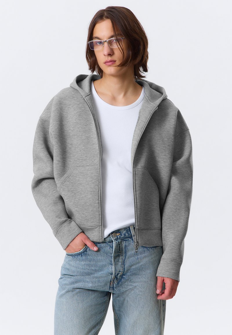 Siva zip-up kapuca s kapuco, izdelana iz mehkega materiala. Ima ribane manšete in dve stranski žepki, nosi se čez belo majico in svetlo modre kavbojke.