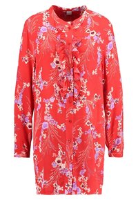 Blouse fleurie rouge à manches longues, avec un détail froncé sur le devant, des boutons au centre et un motif de fleurs violettes et blanches.