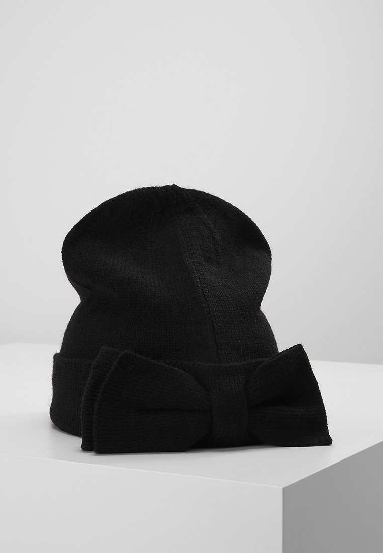 Bonnet en tricot noir avec un large revers et un grand nœud décoratif reposant sur une surface blanche.