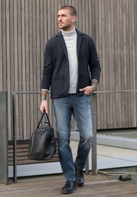 Man in een zwart colbert, lichtgrijze coltrui, blauwe spijkerbroek en zwarte laarzen loopt buiten met een zwarte leren duffeltas.