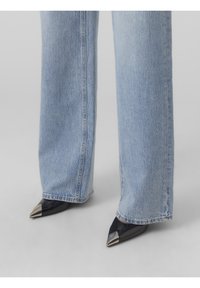 Blå denimjeans med vida ben kombinerade med spetsiga svarta lackklackar med metalliska silverdetaljer på tårna. Mjukt tyg med synliga sömmar.