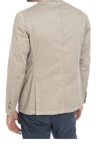 Beige bomullblazer med strukturerade axlar, långa ärmar och två sidofickor. Har guldtonade knappar på ärmsluten och en ventilerad rygg.