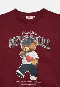 Sudadera color burdeos con un gráfico de un osito de peluche que lleva una camiseta blanca y pantalones azules, con el texto "Estados Unidos NUEVA YORK" arriba y una cita debajo.