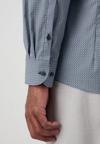 Hellblaues Hemd mit einem kleinen weißen geometrischen Muster, ausgestattet mit Knopfmanschetten und Faltendetails; der Stoff wirkt glatt und leicht.