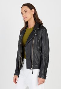 Schwarze Lederbikerjacke mit silbernen Reißverschlüssen, Reverskragen und Reißverschluss-Taschen. Gestylt über einem grünen Oberteil und weißen Hosen, das eine figurbetonte Form zeigt.