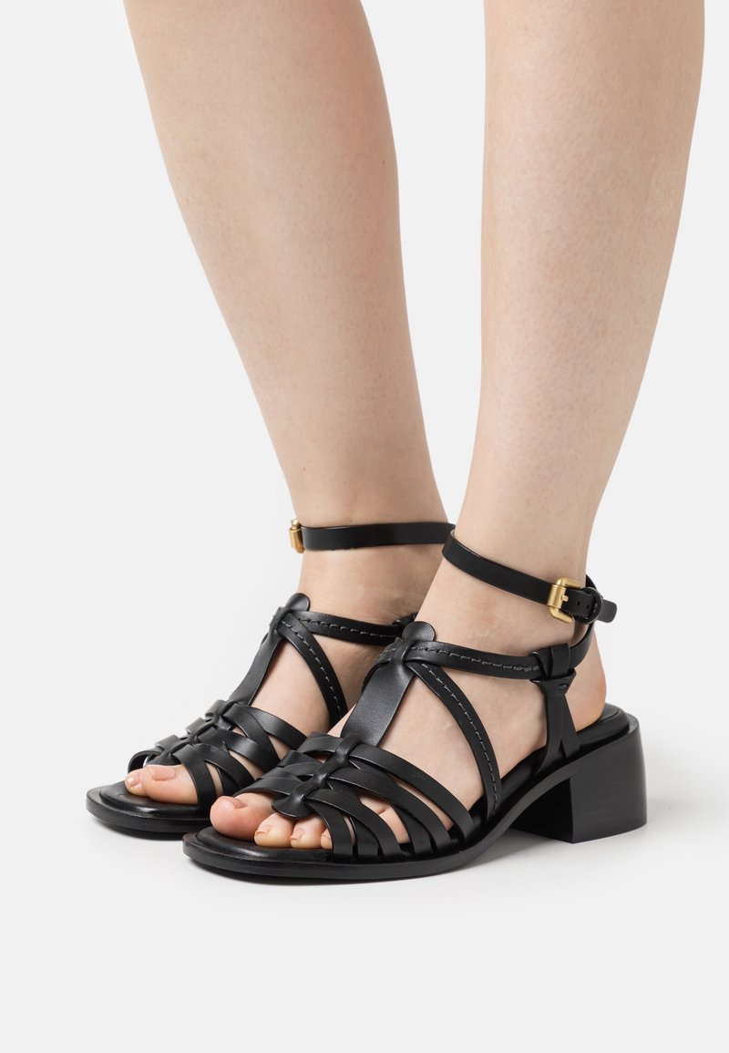 Sandales à talons en cuir noir avec un design tissé, une bride de cheville et un talon bloc rectangulaire. Texture lisse et détails de couture minimaux.
