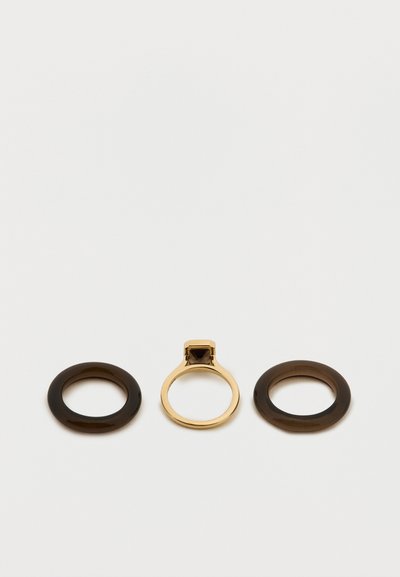 Trois bagues : deux anneaux noirs circulaires et une bague en or avec une pierre carrée foncée au centre. Surfaces lisses et brillantes.