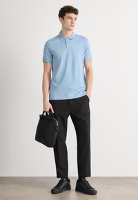 Gaiši zila polo krekls, melni bikses un melni čības. Vīrietis ar melnu somu, stāvot pret neitrālu fona.