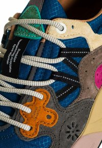 Sneaker multicolore con una tomaia texturizzata blu, accenti in suede marroni e arancioni, lacci bianchi e dettagli di cucitura intricati.