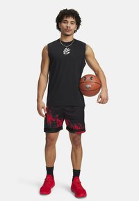 Černé bezrukávové tričko, šortky s vzorem červených a černých plamenů, červené tenisky, držící basketbal s logem Under Armour.
