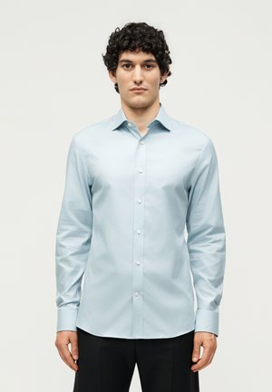 Jeune homme aux cheveux bouclés portant une chemise bleu clair à manches longues et un pantalon noir, debout devant un fond clair uni.