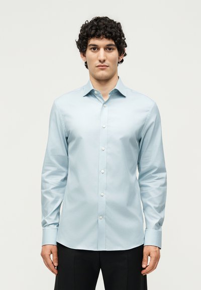 Young man with curly hair wearing light blue long-sleeve button-up shirt and black pants, standing against plain light background.