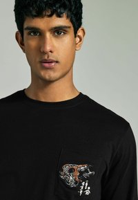 Schwarzes Langarm-T-Shirt mit einer strukturierten Tasche, die ein bunt gesticktes Tigermotiv und japanische Textelemente zeigt.