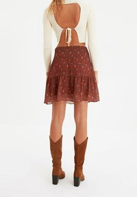 Falda marrón con estampado floral y volantes en capas, combinada con una blusa blanca de manga larga con un gran escote en la espalda, atada en la cintura. Botas marrones hasta la rodilla.