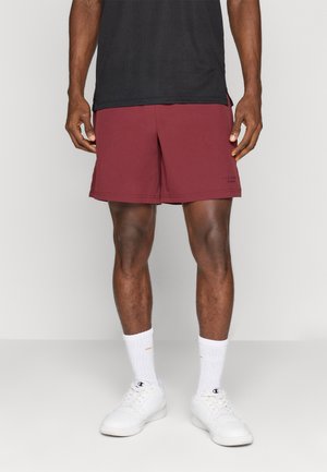 Bordeauxfarbene Sportshorts mit lockerer Passform und strukturierter Stoffoberfläche. Mit einem schwarzen Logodetail auf der unteren linken Seite. Getragen mit weißen Socken und Schuhen.