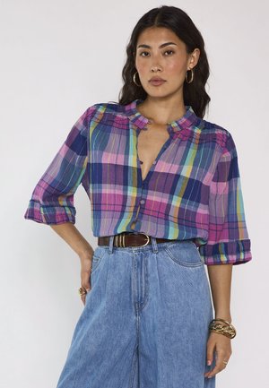 Donna con capelli lunghi e scuri indossa una camicia a quadri colorata con bottoni e jeans a vita alta a gamba larga con cintura marrone su sfondo semplice.