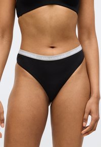 Čierne tangá s vysokým strihom, s sivým elastickým pásom s vyrazeným nápisom "KARL LAGERFELD" v bielych písmenách.