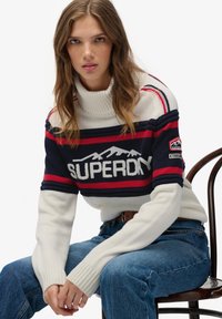 Kobieta siedząca na drewnianym krześle, ubrana w biało-granatowo-czerwony sweter z golfem w paski z logo "Superdry" oraz niebieskie dżinsy.