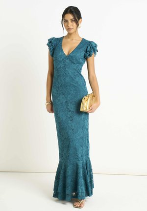 Woman wearing teal lace floor-length dress with ruffled sleeves, holding gold clutch purse, standing against plain white background.