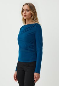 Top lungo a maniche con scollo drappeggiato e dettaglio arricciato sul lato, realizzato in un tessuto liscio e elastico. Abbinato a pantaloni neri.
