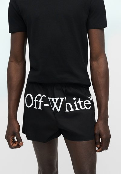 Homme portant un short noir avec un grand logo blanc "Off-White" sur le devant et un t-shirt noir uni à manches courtes.
