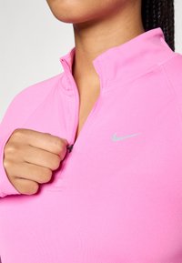 Osoba u svijetloroze Nike majici s četvrtim zatvara�čem, rukom vadi crni zatvarač dolje, otkrivajući vrat i gornji dio prsiju.