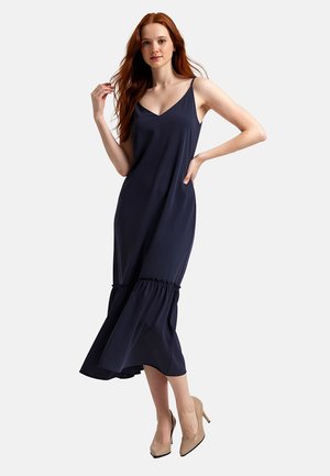 Giovane donna con capelli rossi che indossa un vestito midi senza maniche blu navy e tacchi alti beige, posando con una mano sul fianco.