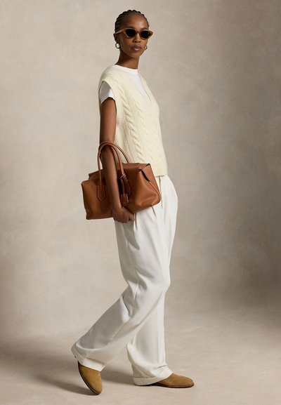 Kobieta ubrana w kremowy sweter bez rękawów z warkoczowym splotem, białe spodnie, beżowe buty, okulary przeciwsłoneczne i niesie dużą brązową skórzaną torebkę.