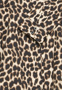 Cămașă cu imprimeu de leopard, având o bază crem și pete maro și negre. Include un nasture frontal și un guler cu două nasturi negri.
