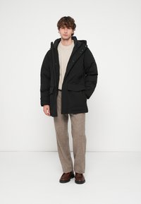 Parka preta acolchoada com fecho de correr, bolsos frontais e capuz, usada sobre um suéter creme em malha trançada e calças bege. Sapatos castanhos completam o visual.