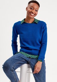 Un pull en tricot bleu avec un motif texturé, superposé à une chemise verte à motifs. L'ensemble est associé à un jean en denim.