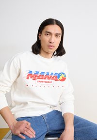 Sweatshirt blanc avec un design graphique coloré comportant des éléments rouges, bleus et jaunes, associé à un jean en denim bleu.
