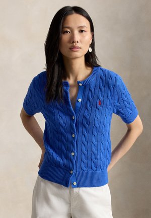 Hosszú sötét hajú nő kék, gombos, rövid ujjú, kábelmintás kardigánban, fehér nadrágban és fehér fülbevalóval, kezét csípőjén tartva áll.