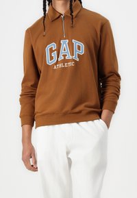 Felpa con zip marrone con logo GAP Athletic in blu e bianco. Maniche lunghe con polsini a coste, indossata con pantaloni bianchi.