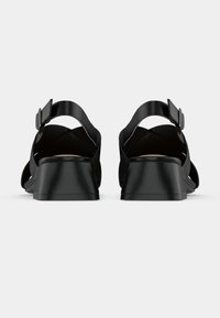 Μαύρα σανδάλια με ανοιχτή φτέρνα, ρυθμιζόμενα λουριά στον αστράγαλο και χαμηλό τακούνι, εμφανιζόμενα από πίσω σε λευκό φόντο.