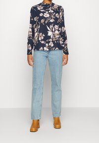 Blusa floreale in blu navy con fiori rosa e beige, con colletto alto e maniche lunghe, abbinata a jeans a gamba dritta azzurri chiari e stivali con tacco color beige.
