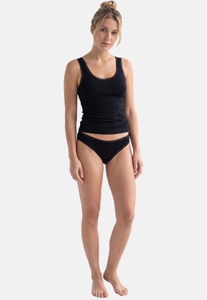 Nainen seisoo paljain jaloin, päällään musta pitsireunainen toppi ja siihen sopivat mustat pitsireunaiset alusvaatteet yksinkertaisella vaalealla taustalla.